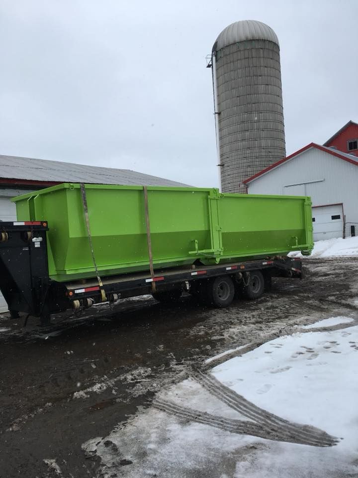 Benne verte sur une remorque, stationnée sur un sol enneigé près d'un silo et d'un bâtiment au toit rouge.