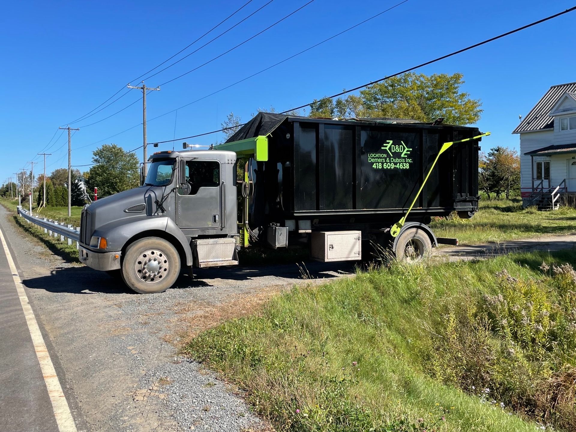 Camion à benne basculante gris stationné sur un accotement en gravier à côté d'une route, avec un grand conteneur noir.