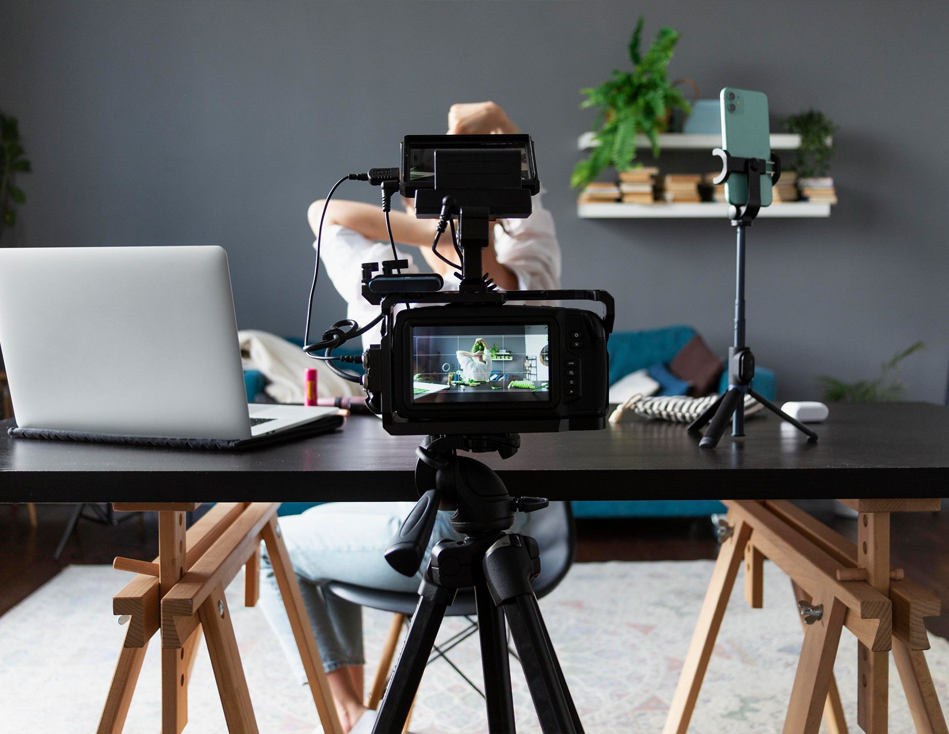 Person setting up video recording equipment at desk; laptop, cameras, and phone on tripod.
