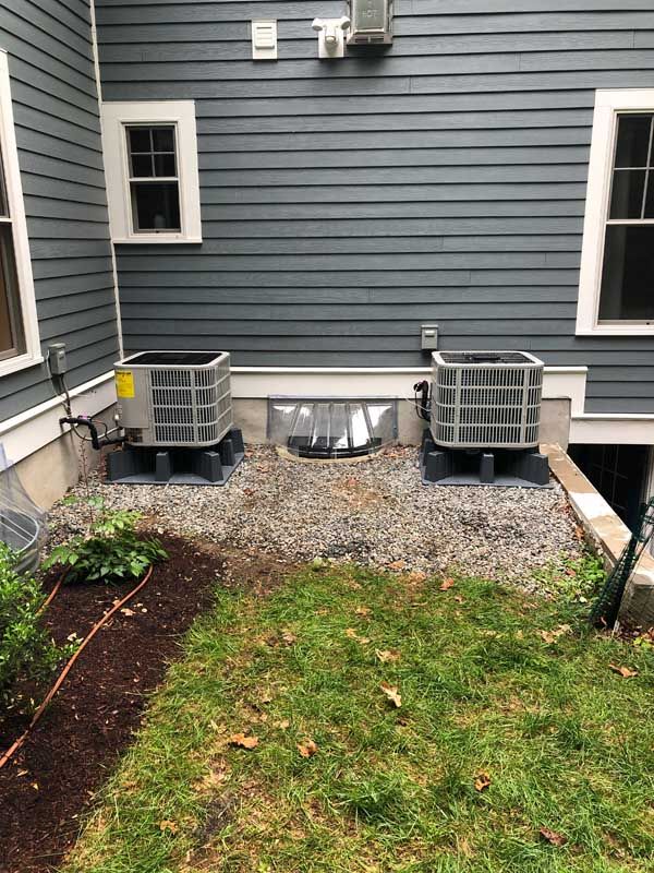 Two air conditioning units on gravel by a gray house.
