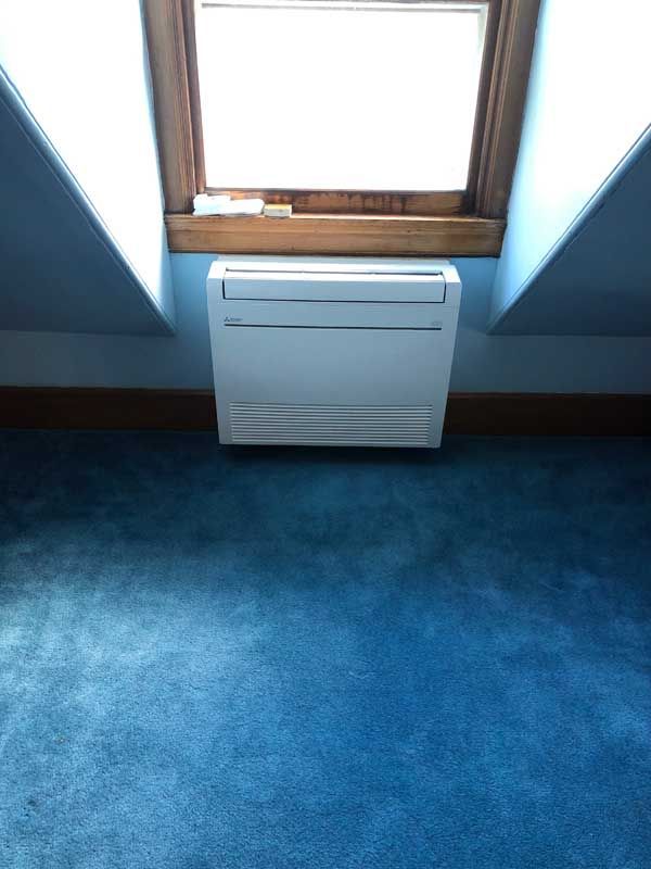 White air conditioner under a window in a room with blue walls and carpet.