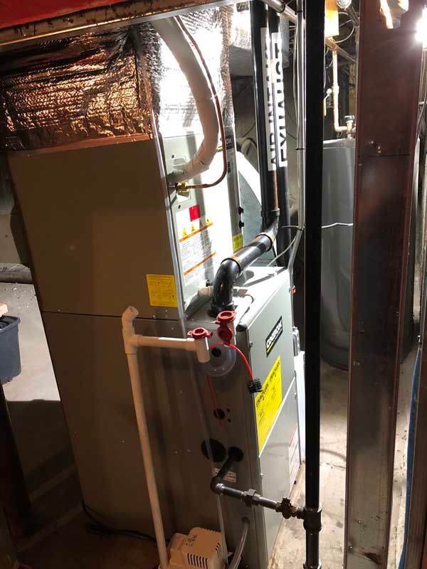 Furnace with ductwork in a basement setting. Silver and gray mechanical unit, pipes, and insulation visible.