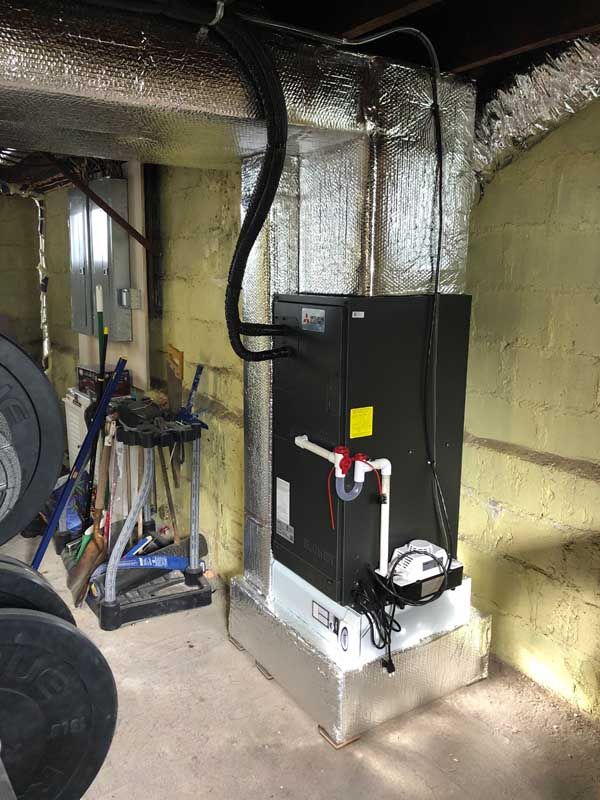Black HVAC unit in a basement setting. Silver insulation, gray pipes, and concrete walls are visible.