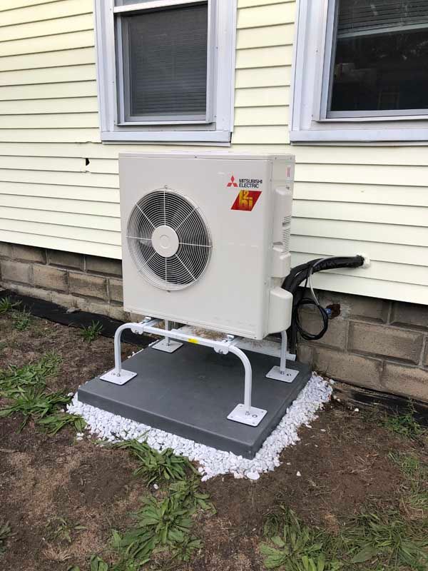 Mitsubishi heat pump unit on a concrete pad next to a yellow house with windows, supported by metal legs.