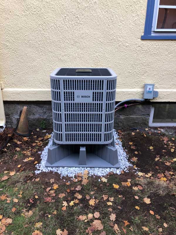 Gray air conditioning unit on a gray base, set on gravel near a yellow house with a window and fall leaves.