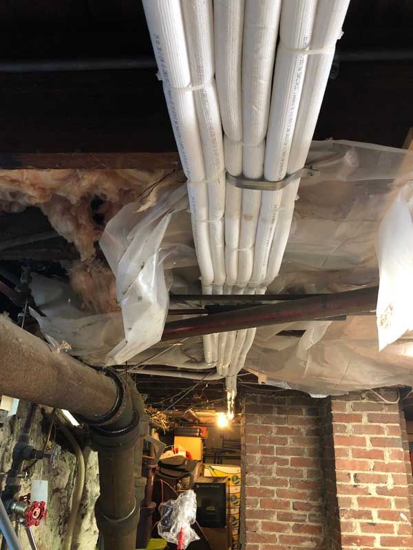 White PVC pipes bundled together, suspended in a basement near brick wall and other pipes.