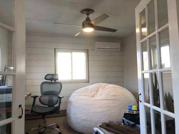 A small office with a desk, chair, beanbag, and air conditioner; bright with natural light.