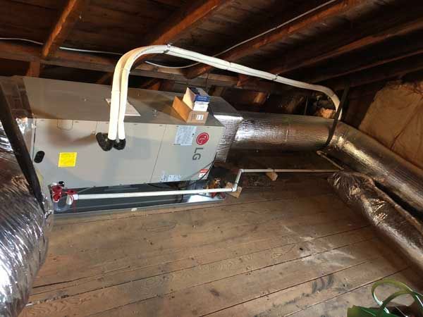 HVAC unit in an attic with ductwork. Metal unit, silver insulated ducting, wood boards.