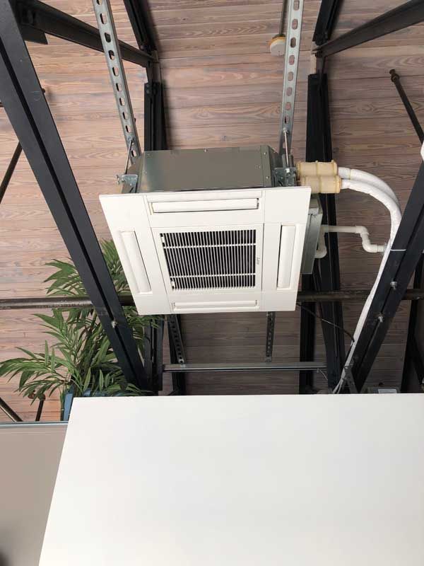 Ceiling-mounted air conditioner with a white panel, suspended from metal supports. White pipes extend from the unit.