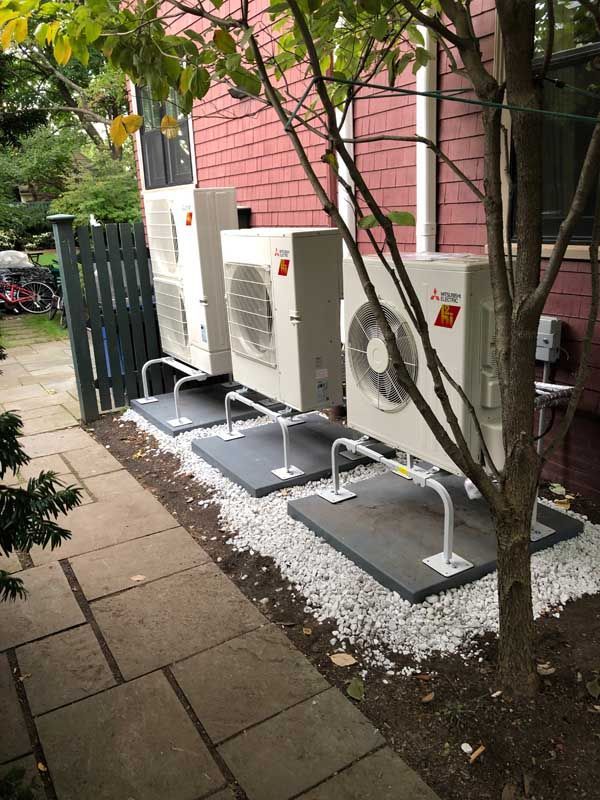 Three Mitsubishi air conditioning units on gray pads next to a red building and a gray fence.