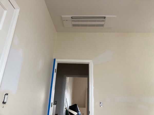 Ceiling-mounted HVAC unit above doorway in room with off-white walls, doorway taped blue.