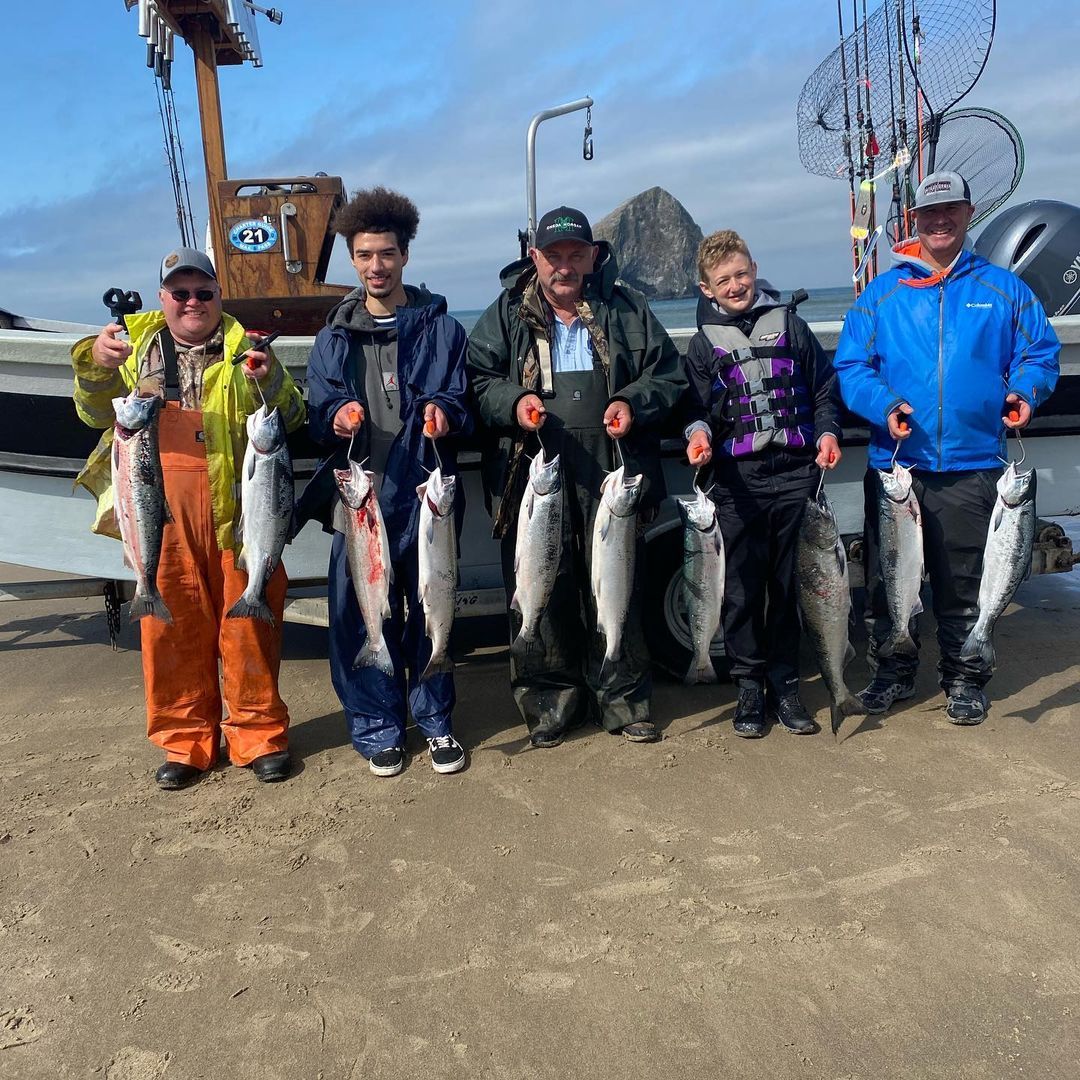 Five people proudly display their freshly caught salmon near a boat on a coast, sunny day.