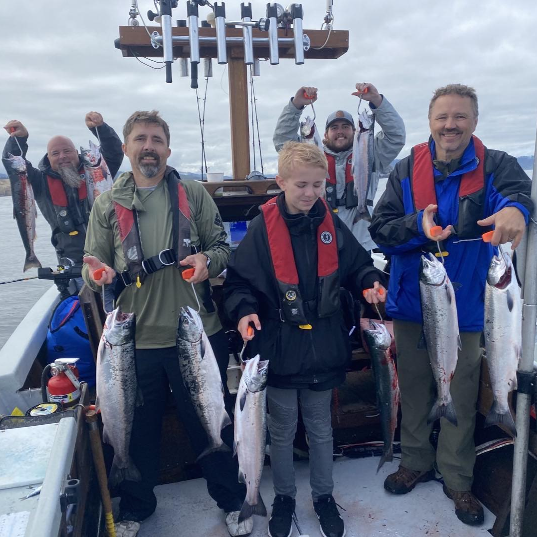 Group of people on a boat, smiling and holding up freshly caught silver fish. Cloudy sky, nautical setting.