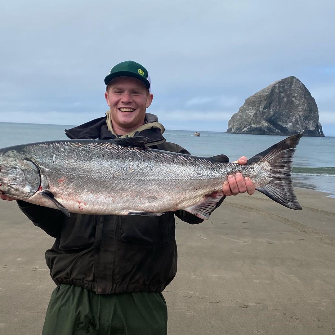 Man smiles, holding a large silver salmon on a beach with a rocky island in the background.