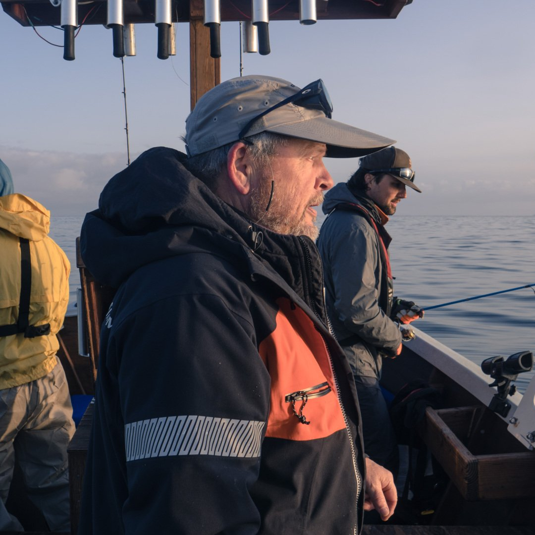 Man on a boat fishing, wearing a hat and jacket, looking at the ocean. Two others in the background.