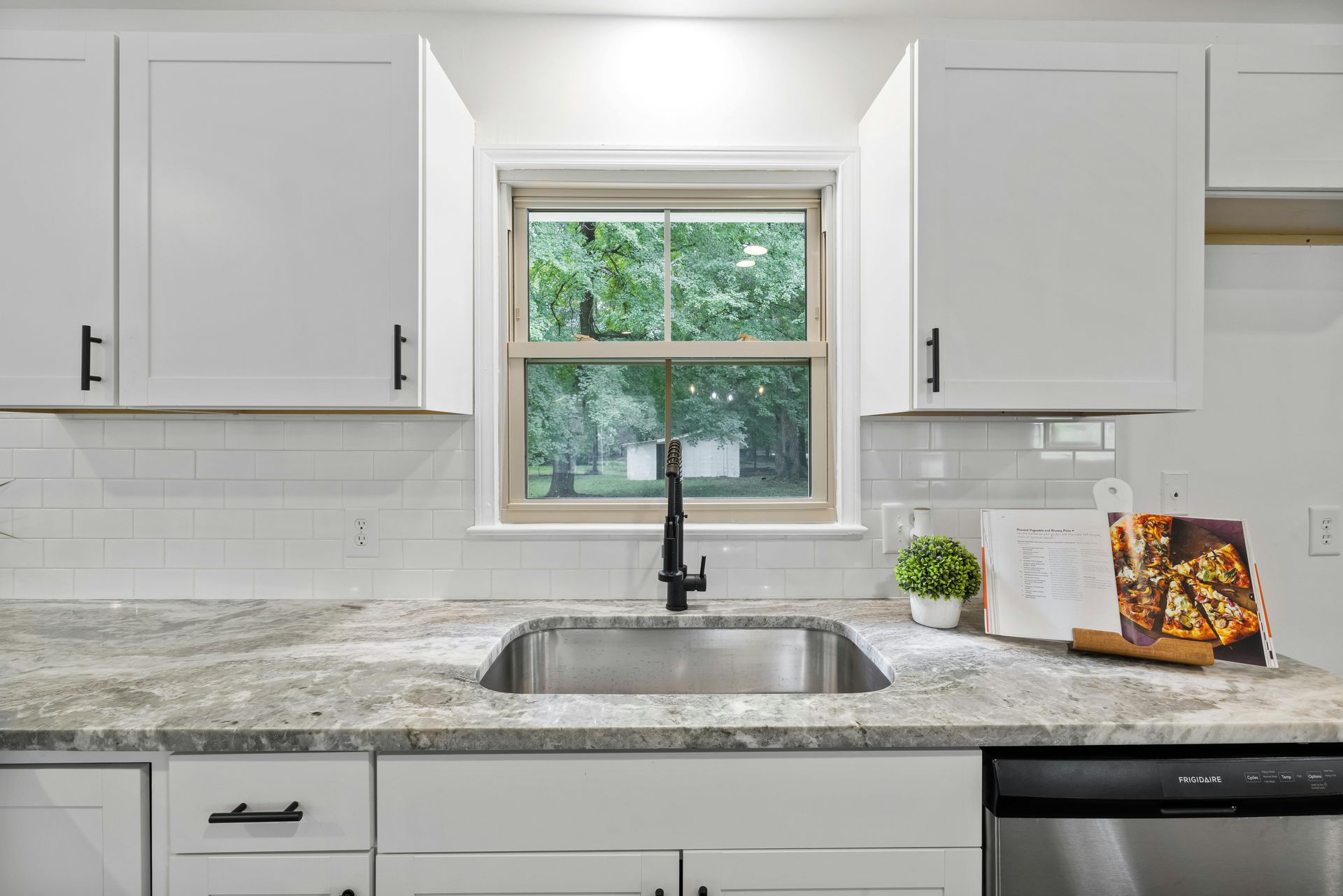White kitchen with a stainless steel sink, countertop, and cabinets. A window looks out to green trees.