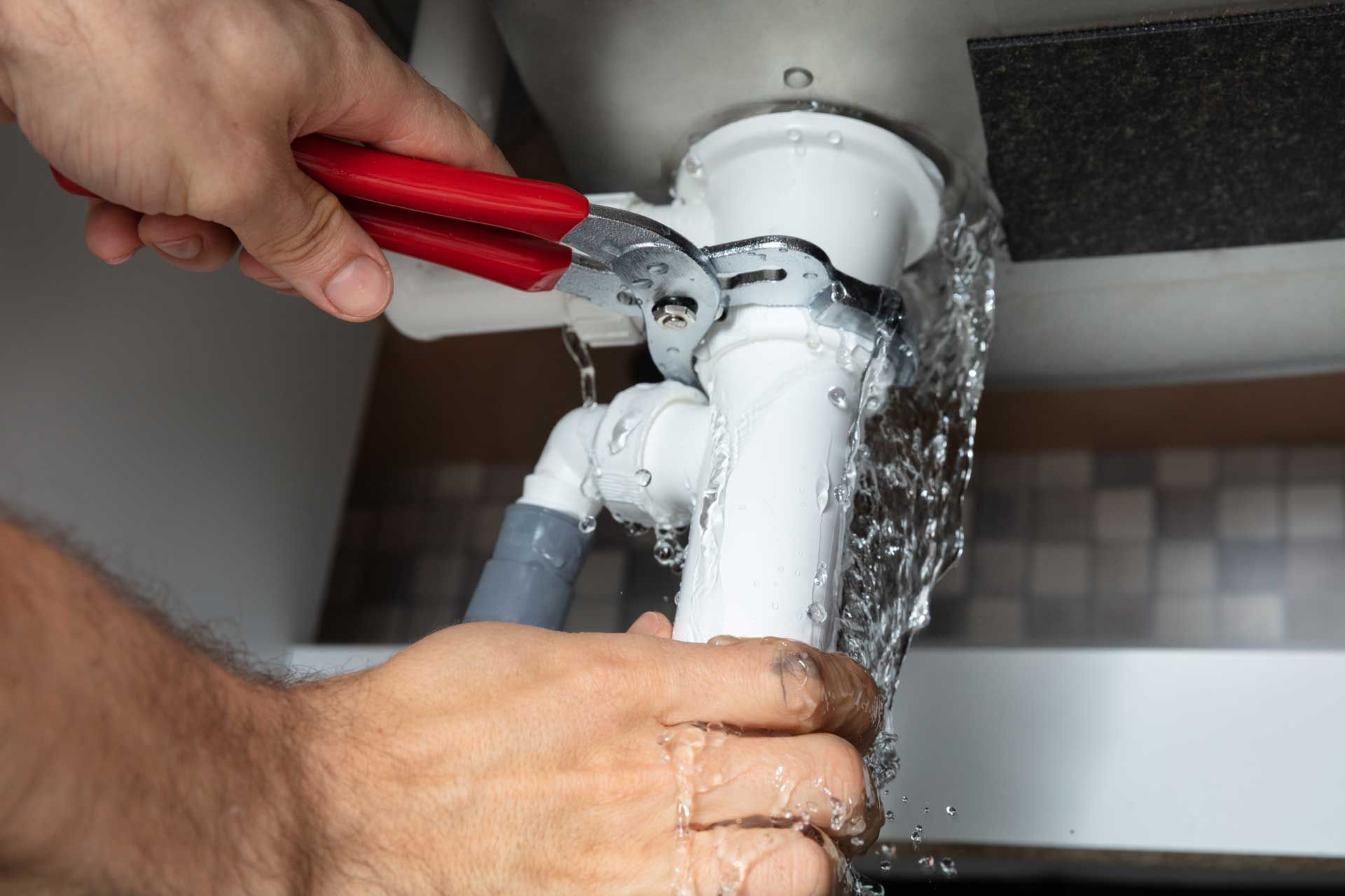 Hands using pliers to tighten a leaking white pipe under a sink, with water spraying out