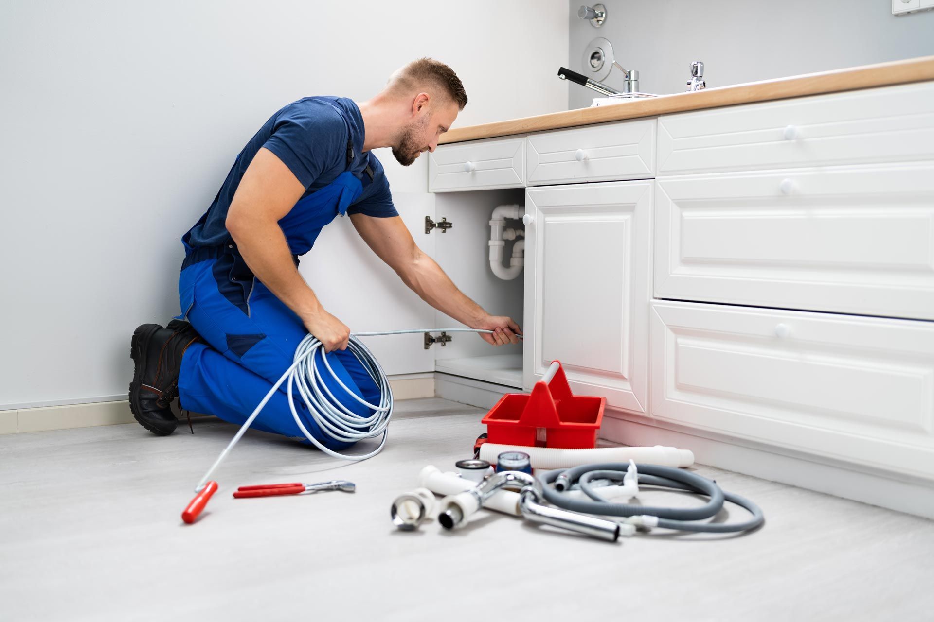 Plumber in blue uniform repairing pipes under a white kitchen sink with tools on the floor