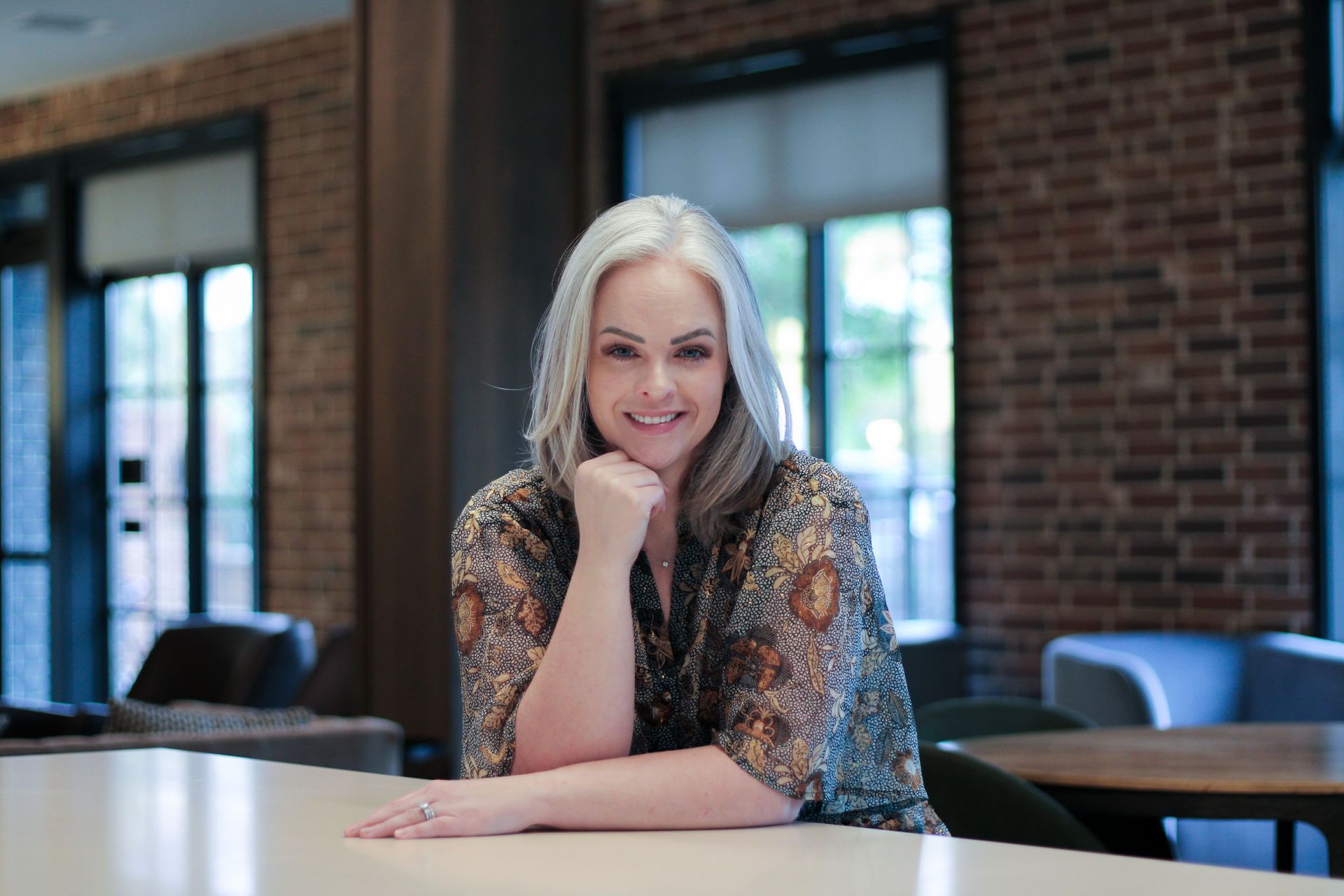 A woman is sitting at a table with her hand on her chin and smiling.