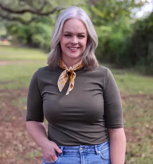 A woman wearing a green shirt and blue jeans is standing in a field.