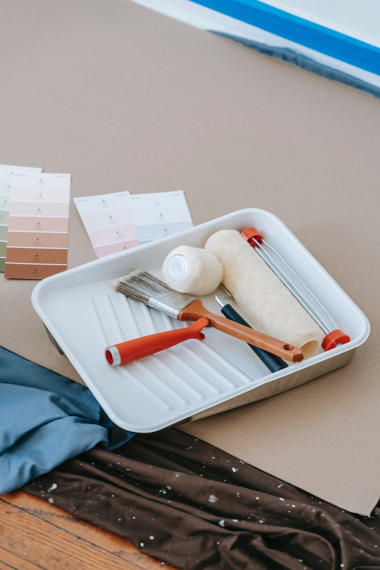 A tray with paint rollers , brushes , and paint samples on a wooden floor.