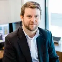 A man with a beard is wearing a suit and sitting in front of a window.