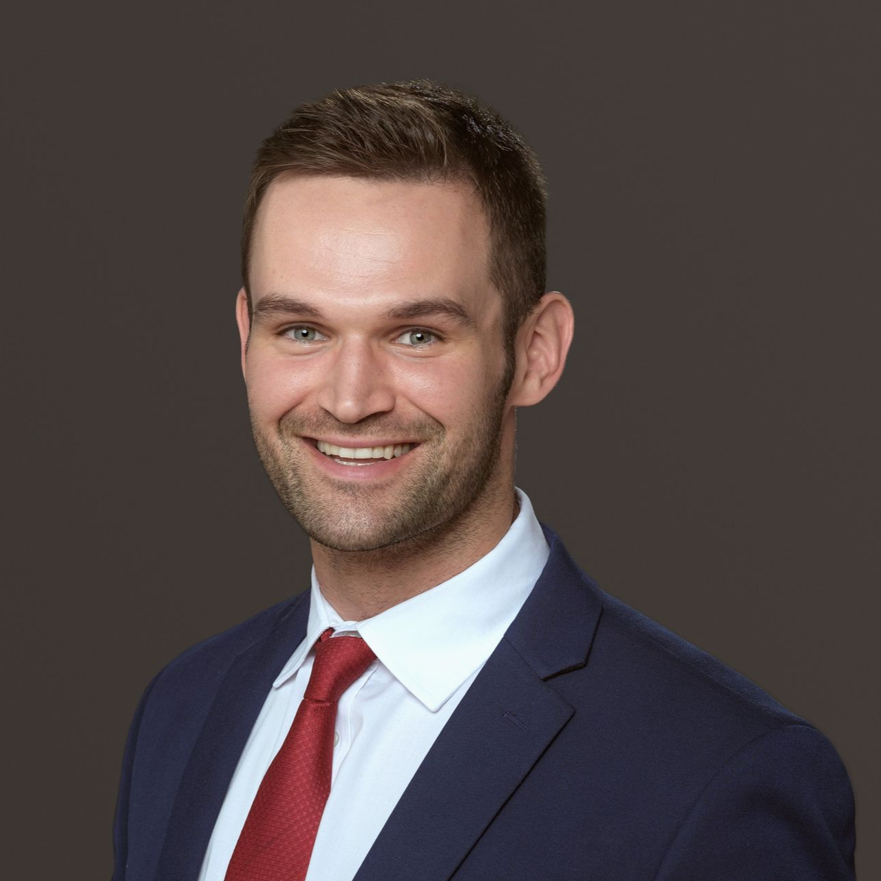 Man in a blue suit and red tie smiling, with short brown hair, against a dark gray background.