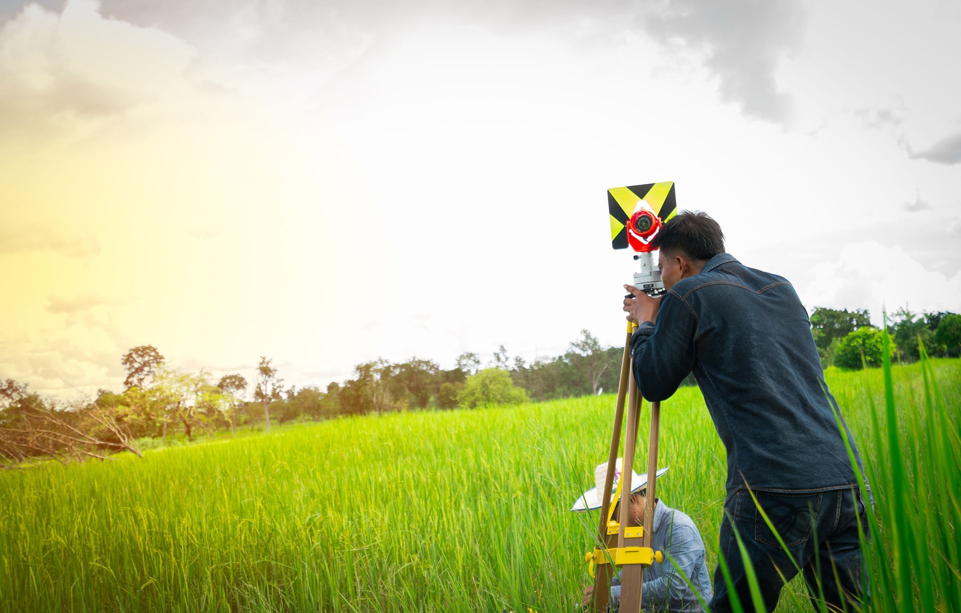 Surveyor using equipment to measure land in a grassy field on a cloudy day.