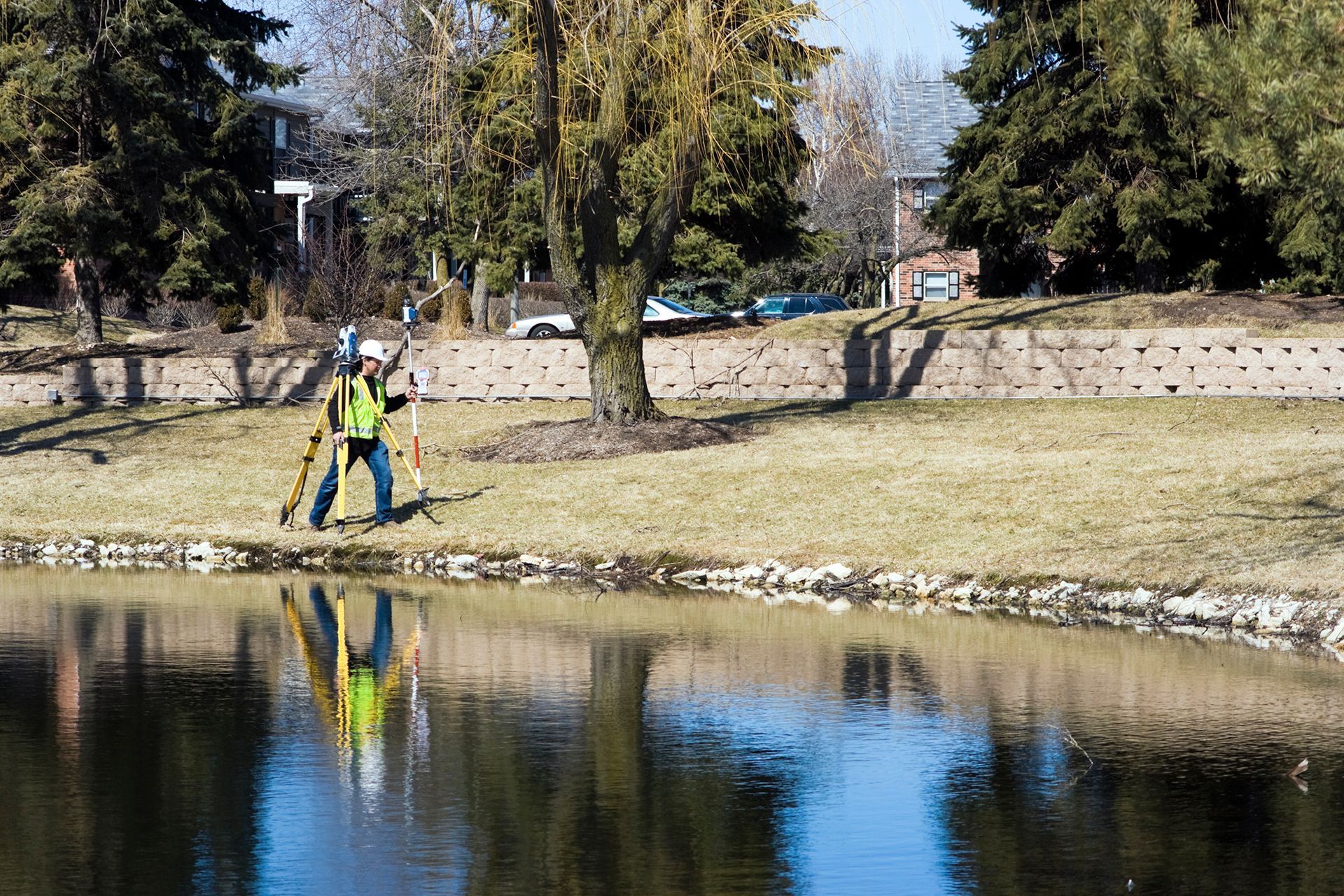 Surveyor in safety vest using equipment by a body of water, reflected in the water, with trees and buildings in the background.