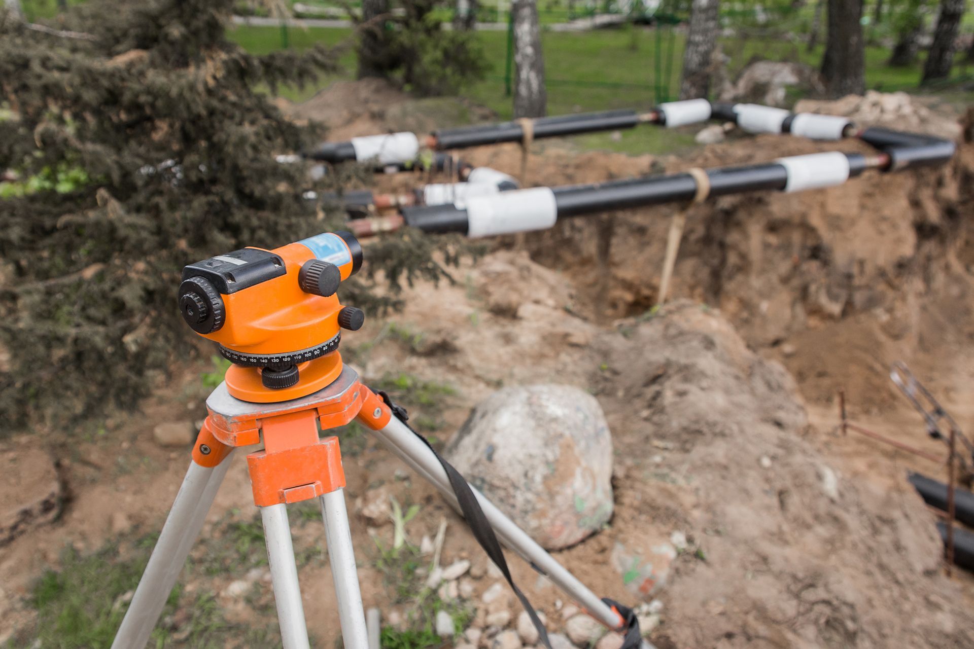 Orange surveying level on a tripod, in front of exposed pipes, outdoors.