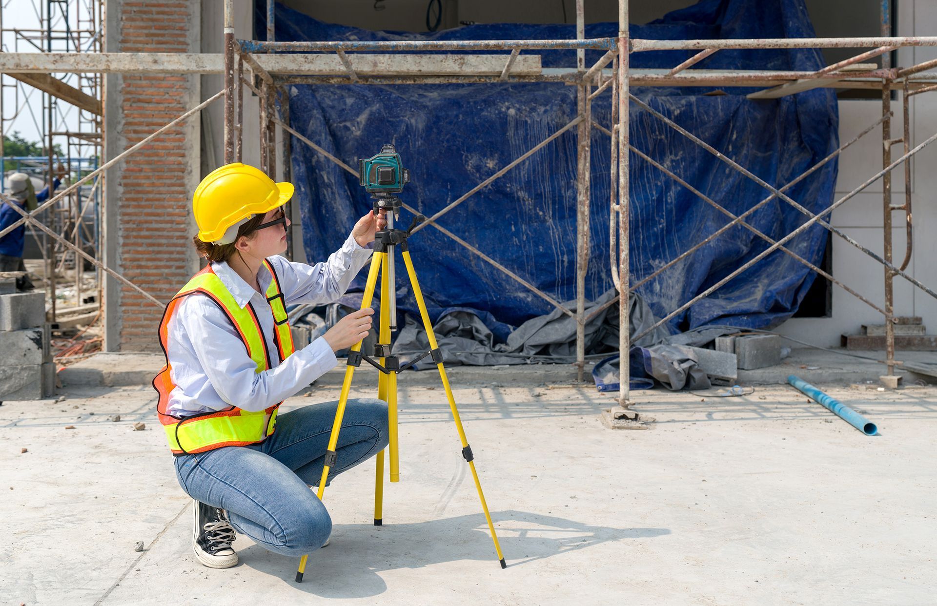 Woman in safety vest and hard hat using a surveying level on a construction site.