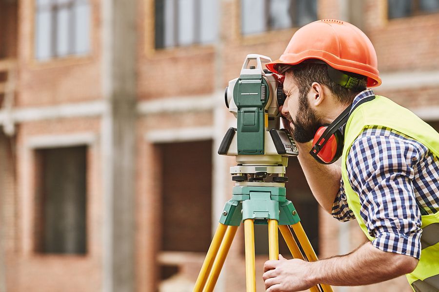 Surveyor in a hard hat and vest looking through a theodolite on a tripod at a construction site.