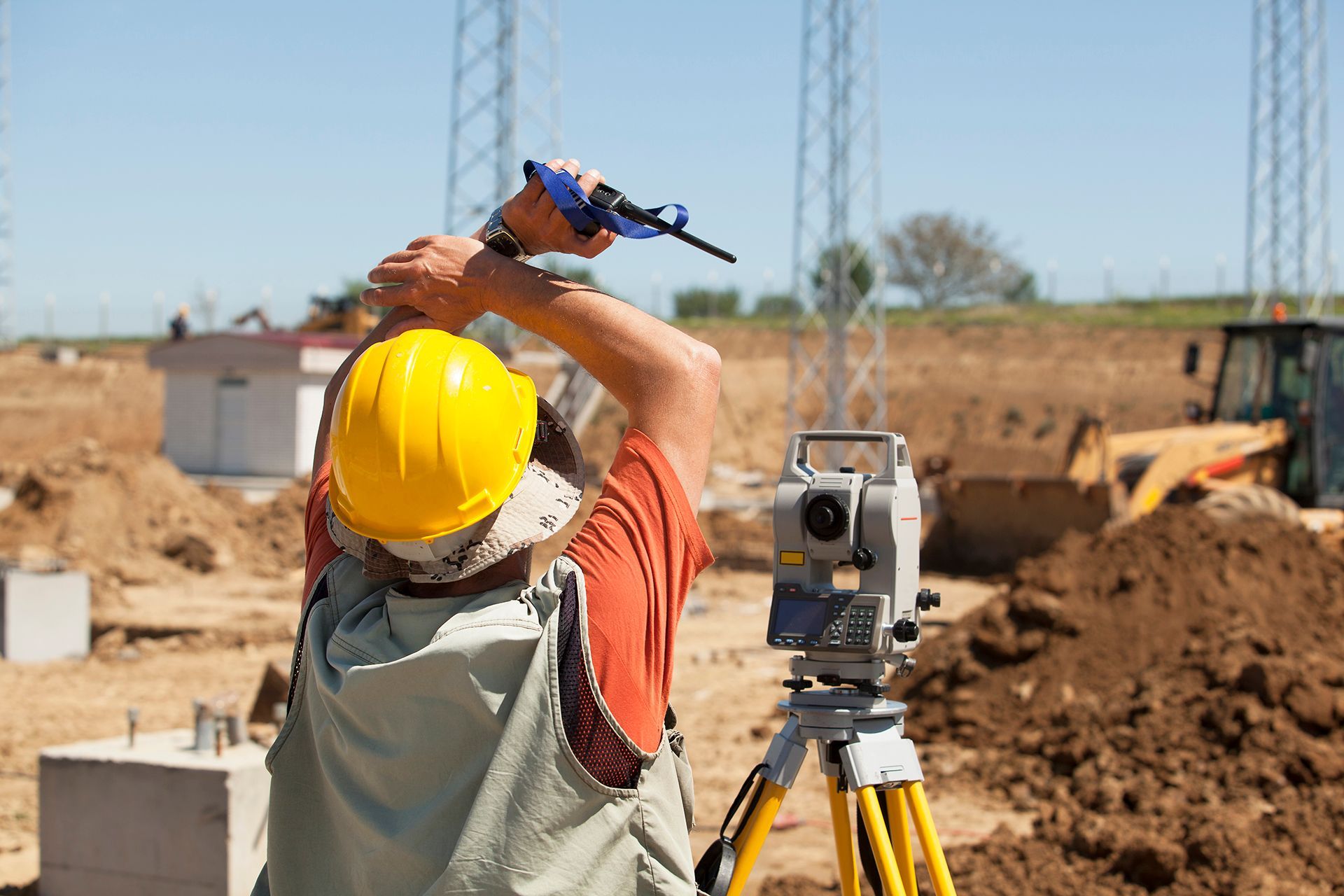 Construction worker in a yellow hard hat uses surveying equipment on a construction site.