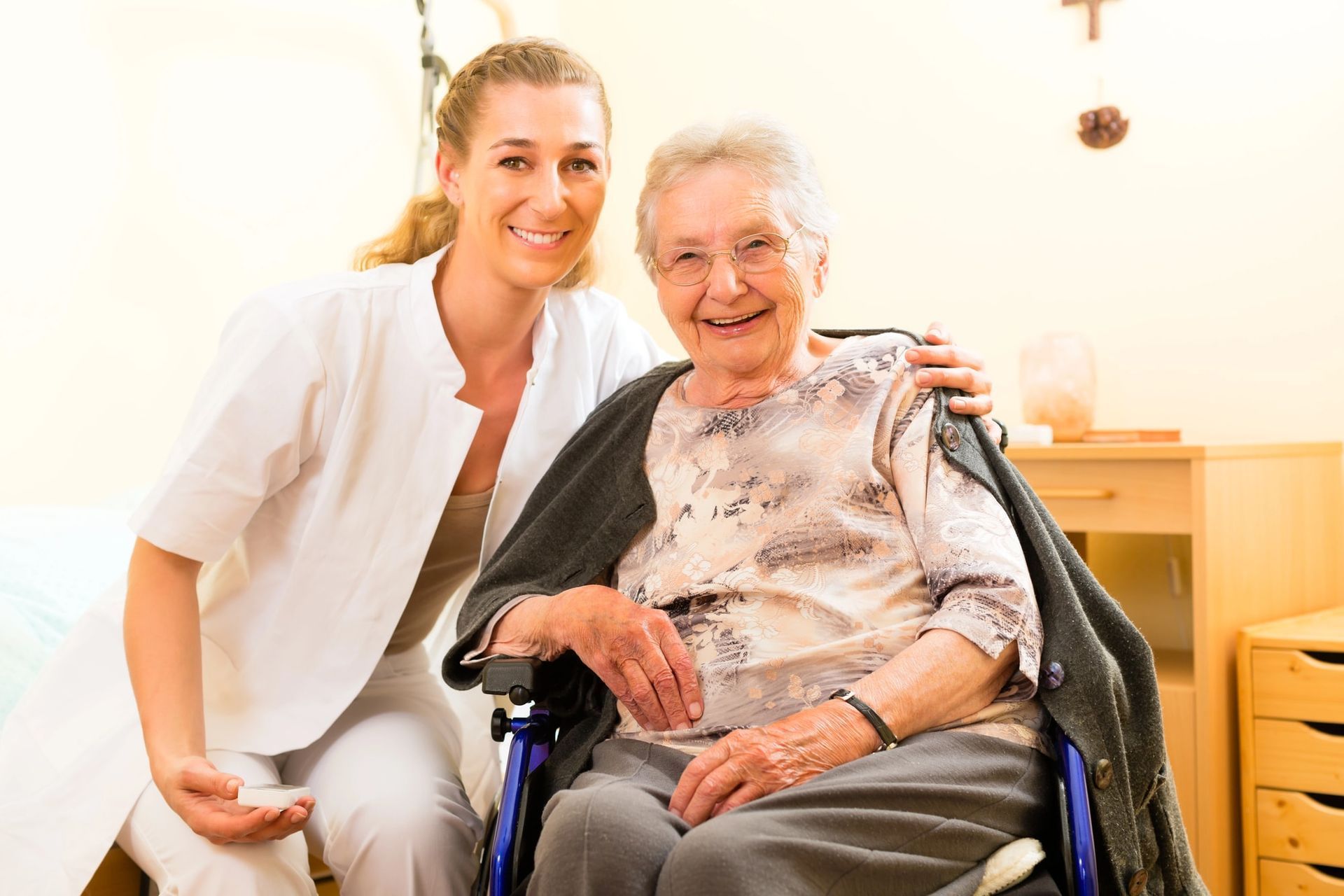 Woman in scrubs smiles, arm around elderly woman in wheelchair, both smiling, in a bright room.