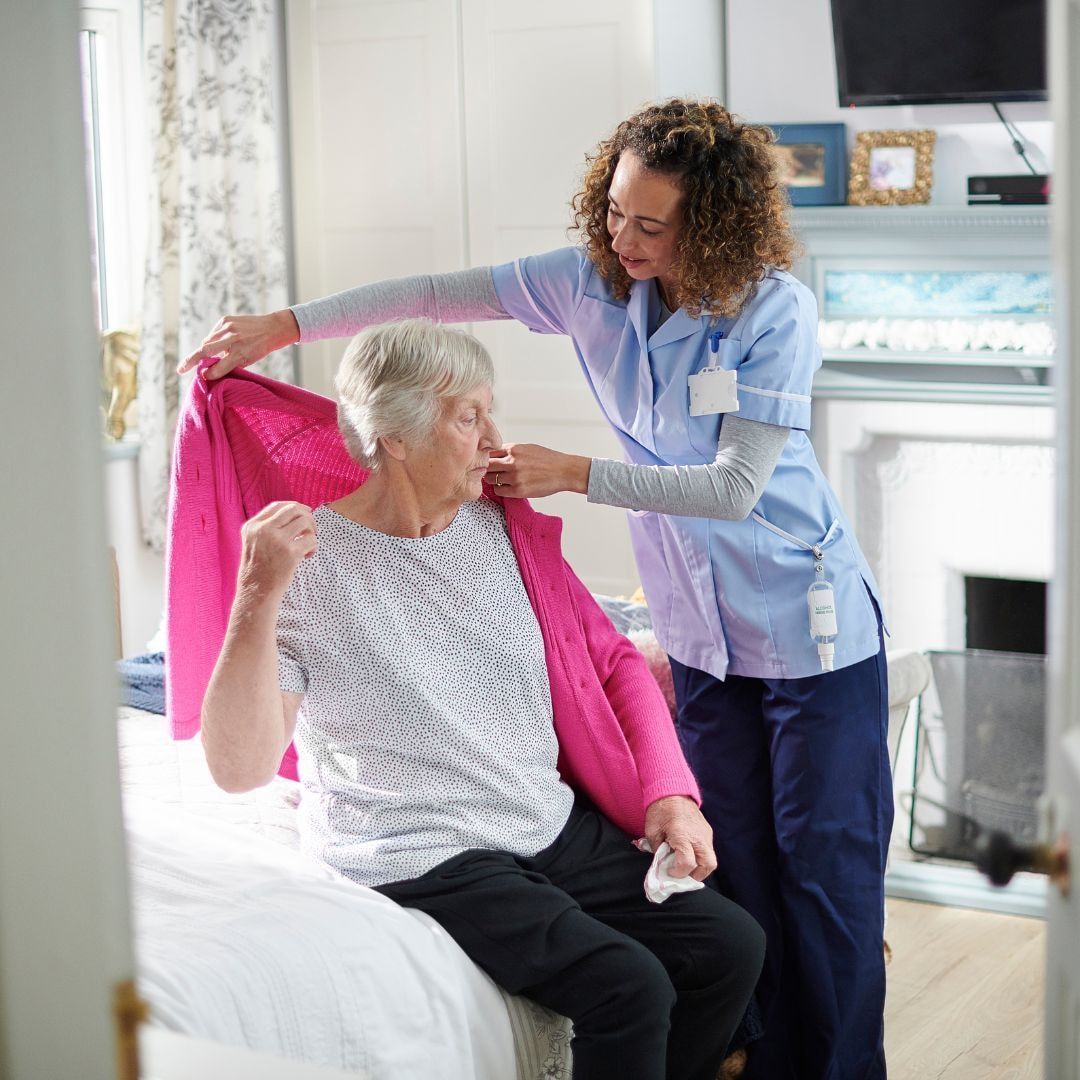Caregiver helping senior put on a pink sweater in a bedroom.