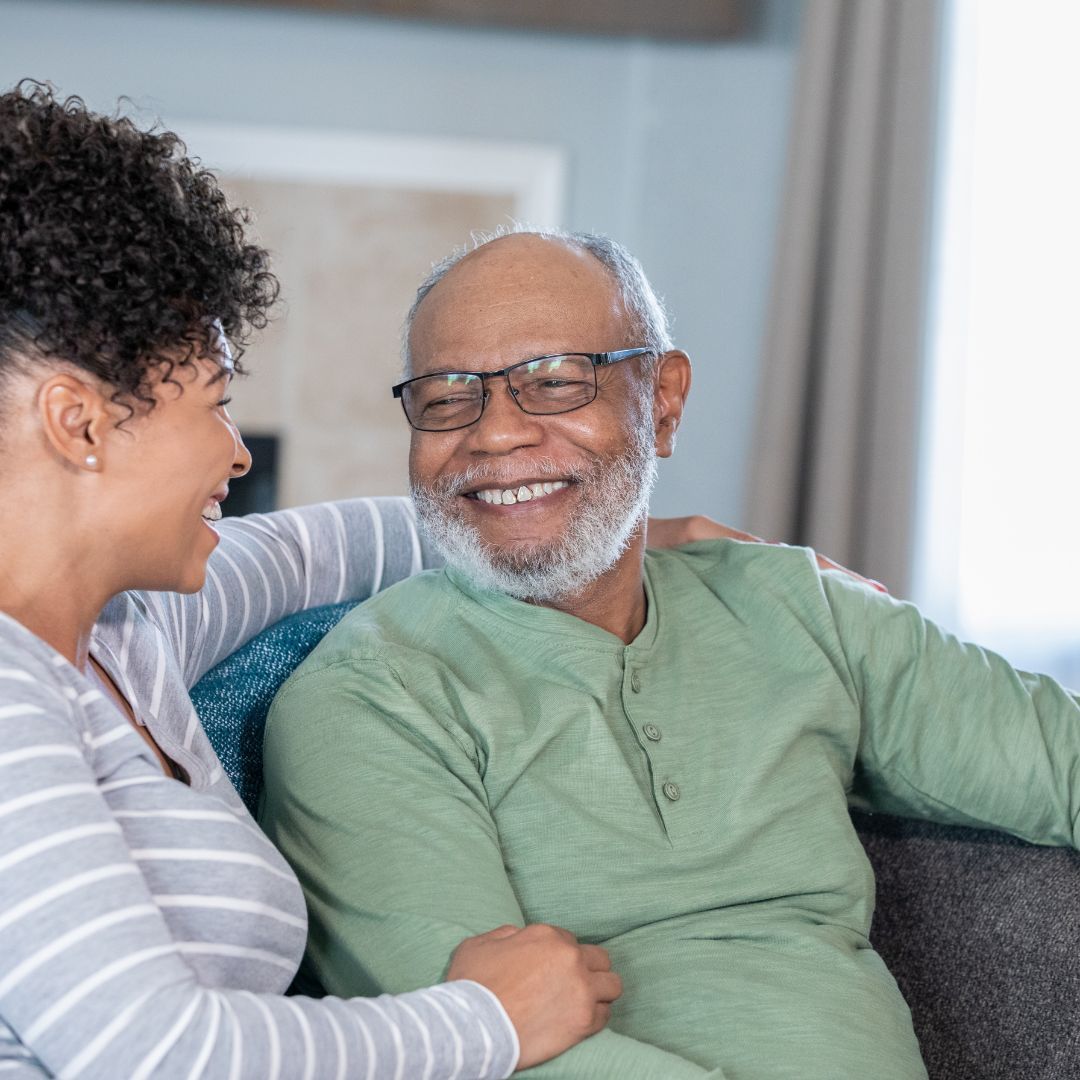 A smiling person in a green shirt sits with a person in a striped shirt on a couch.