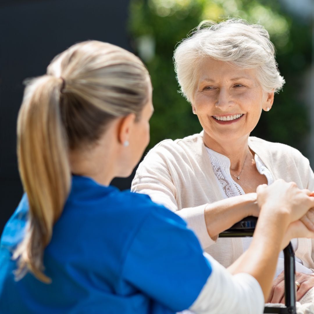 Caregiver in blue scrubs speaking with smiling elderly woman outdoors.