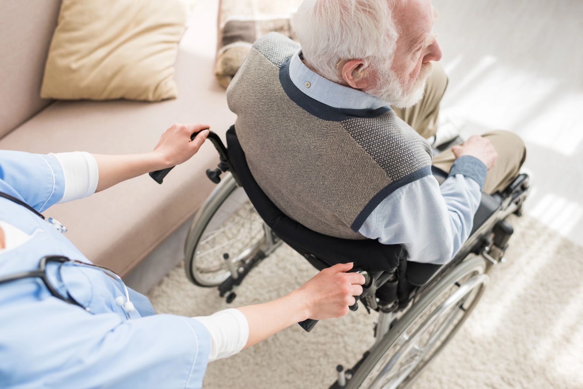 A caregiver pushing a person in a wheelchair indoors near a sofa.