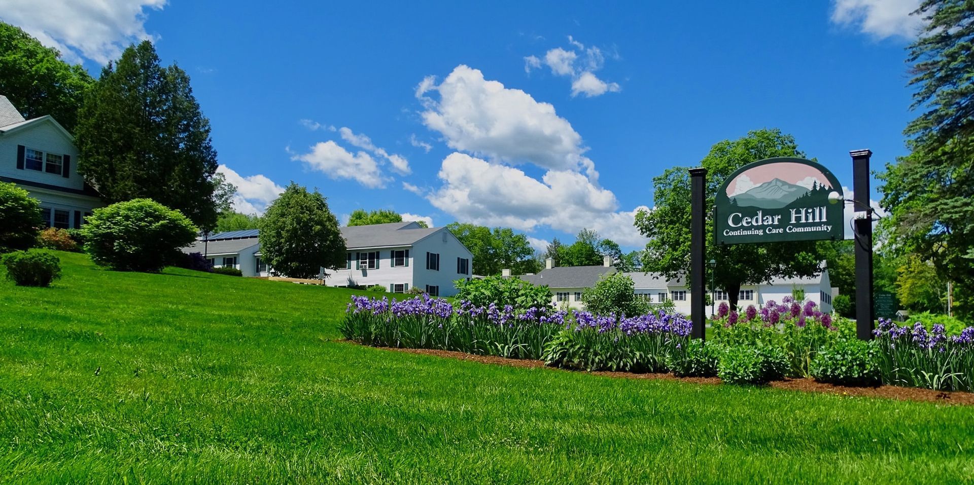 Green lawn with white buildings, sign for Cedar Hill under a blue sky with clouds.