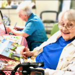 Two older adults painting at a table, one smiles at the camera, art supplies visible.