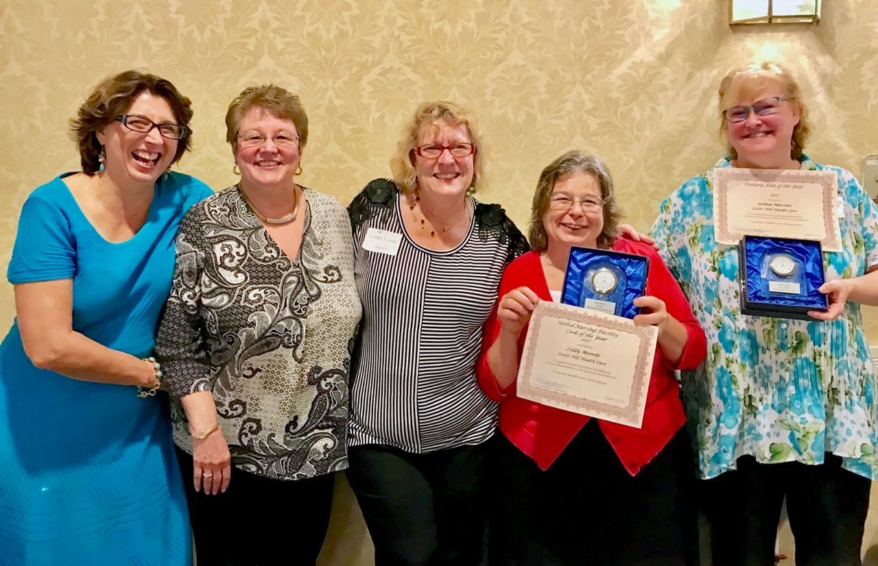 Five women smiling, holding awards. Indoors with patterned wallpaper.