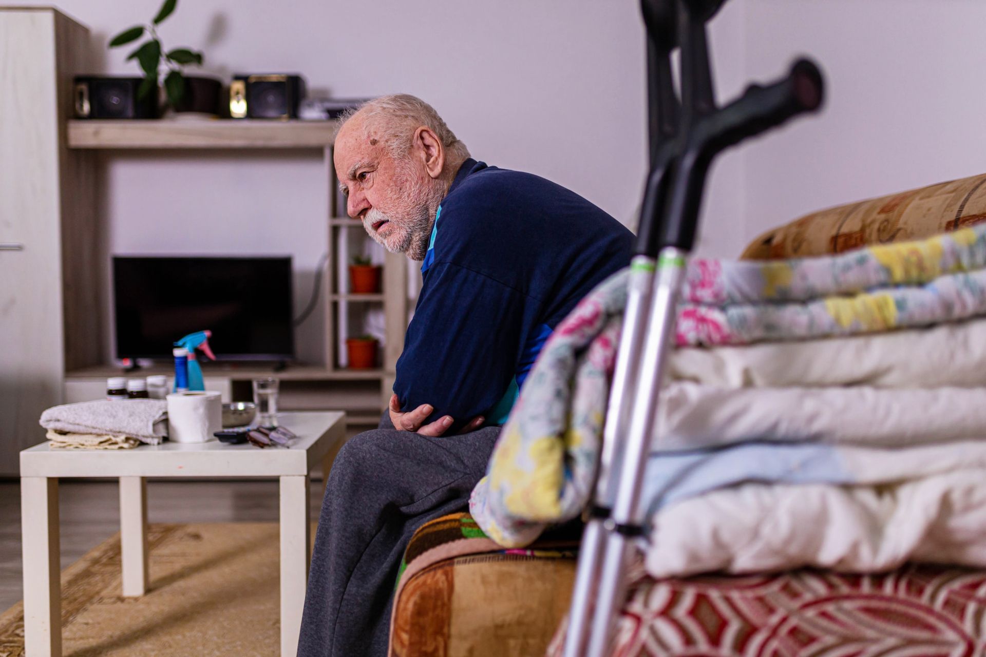 Elderly person sitting on a sofa, arms crossed, looking down, with a crutch and folded blankets nearby.