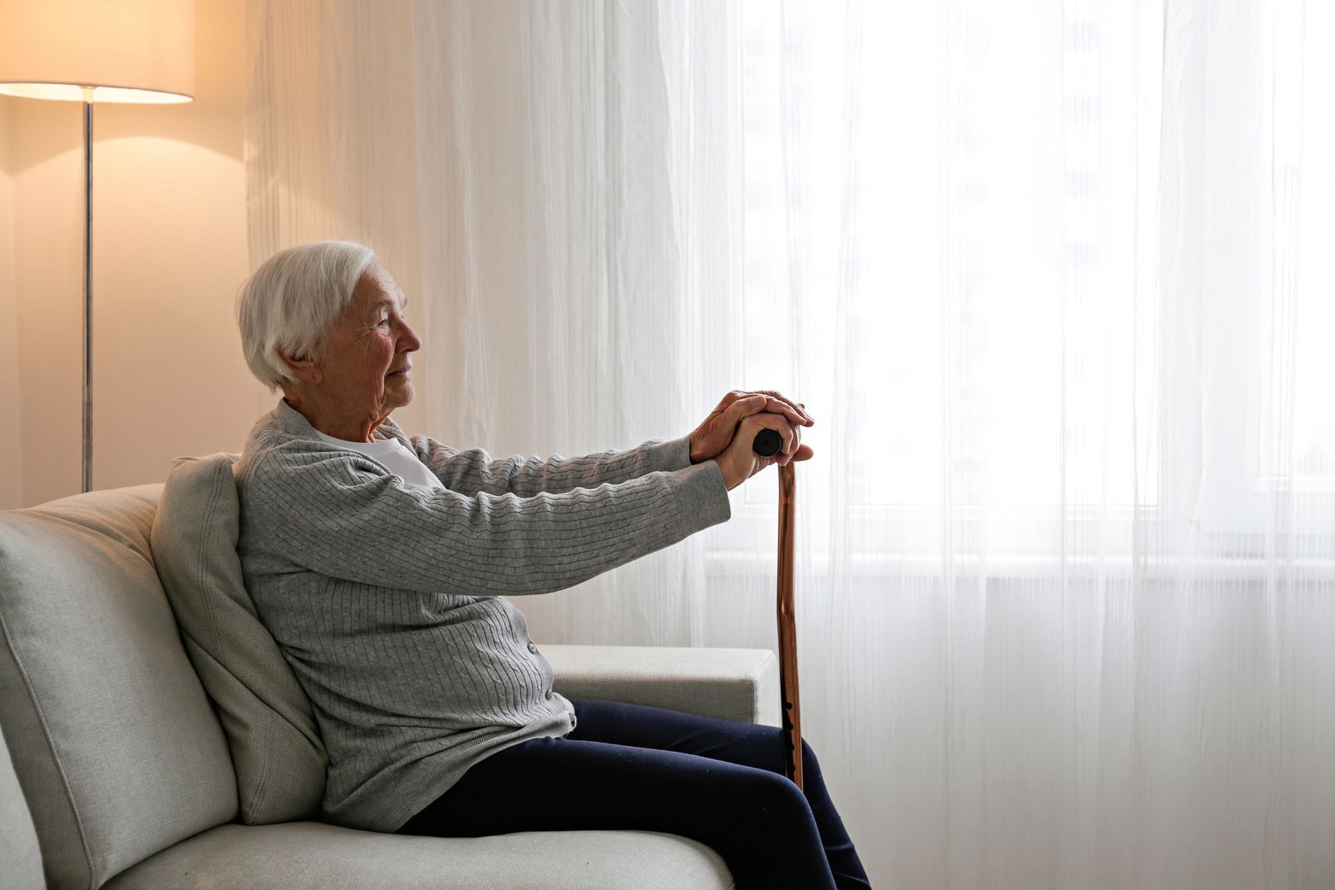 Older person sits on a sofa, holding a cane, looking out a window.