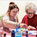 Woman helps another paint at a table with paint supplies. Both smile and look at artwork.