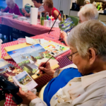 Senior painting class at a table with red gingham tablecloth. Woman paints a landscape while looking at a reference photo.