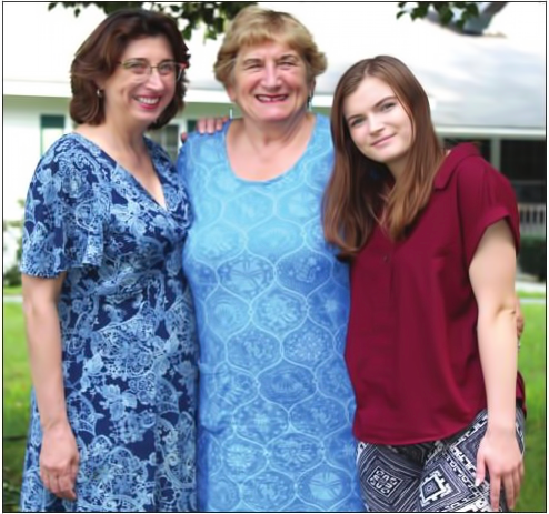 Three women, likely family, smiling outdoors; one in blue dress, one in a red top, one in patterned pants.