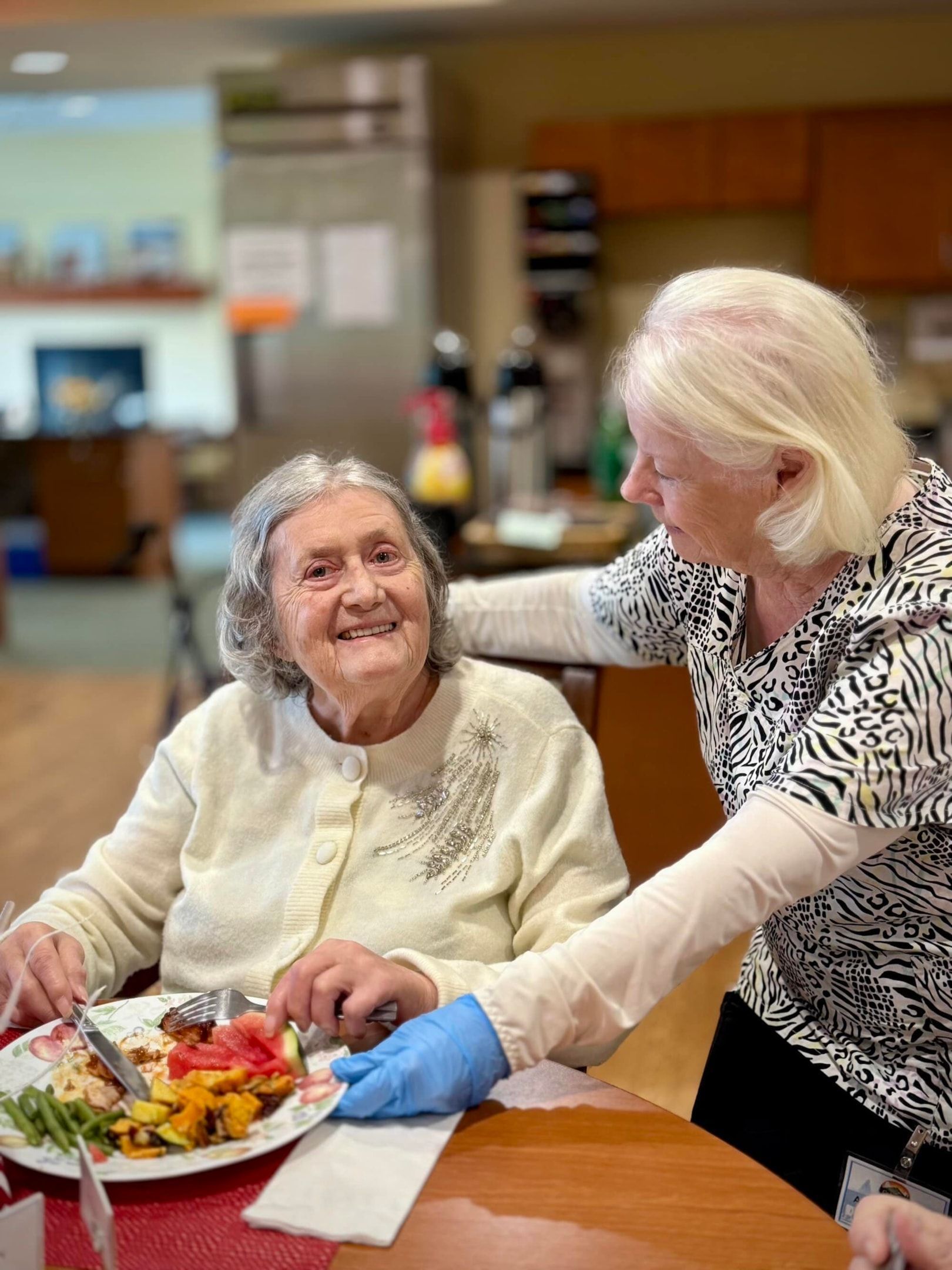 Elderly woman seated at a table, smiling while eating. Caregiver assists her with the meal in a dining room.