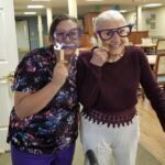 Woman and elderly woman with purple glasses props, smiling in a room.