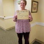 Woman in mauve sweater and maroon pants holds a sign that reads 