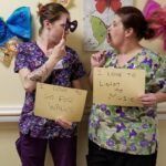 Two women in scrubs holding signs, one with a paper mustache, posing for a photo in a decorated room.